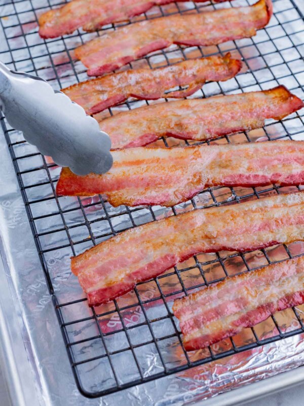 Bacon cooked on a wire rack in a baking sheet is picked up with tongs.