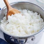 A wooden spoon stirs cooked white rice in a pot.