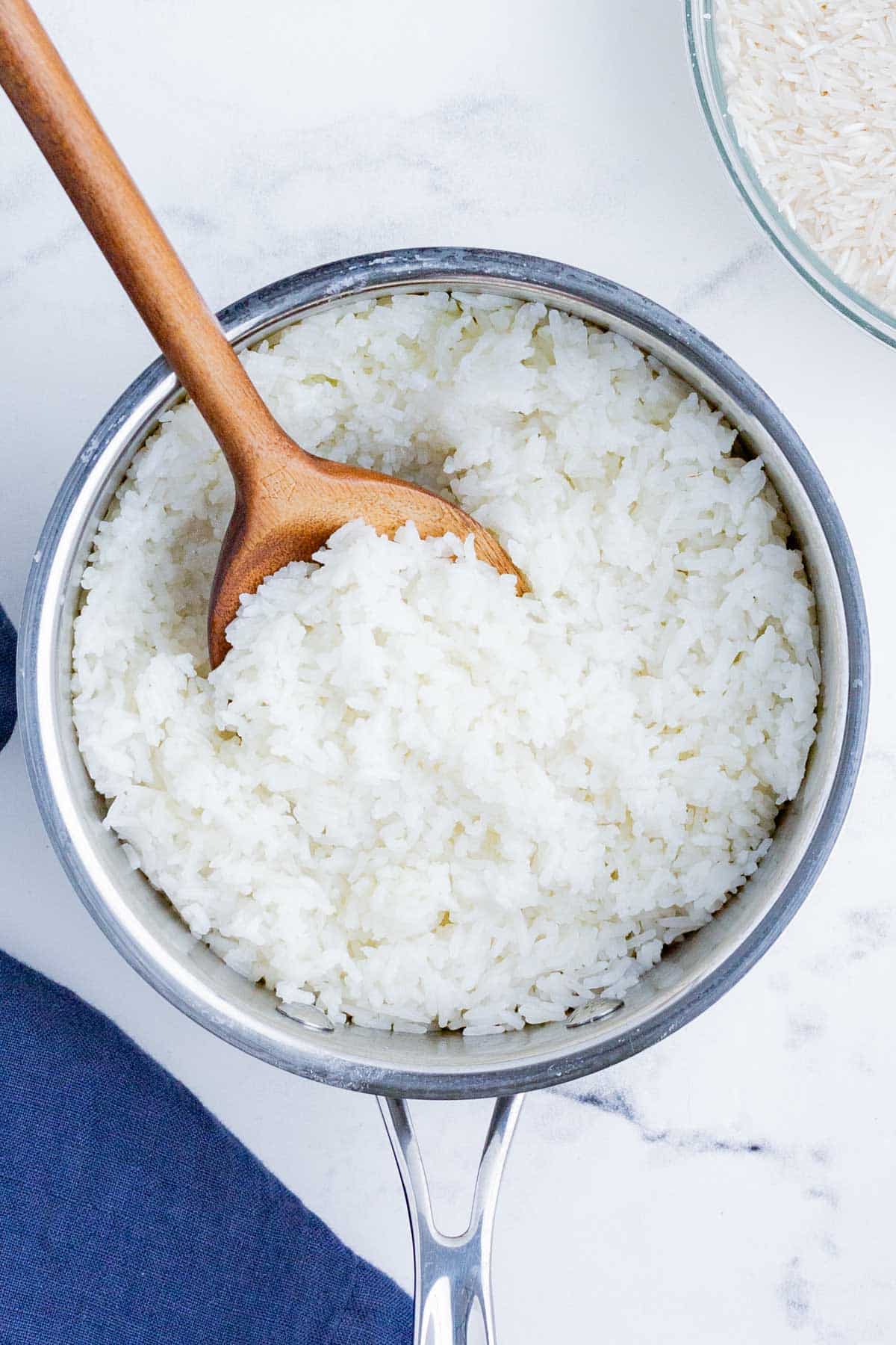 A silver pot full of white cooked rice and a wooden spoon with uncooked rice in a clear bowl on the side.