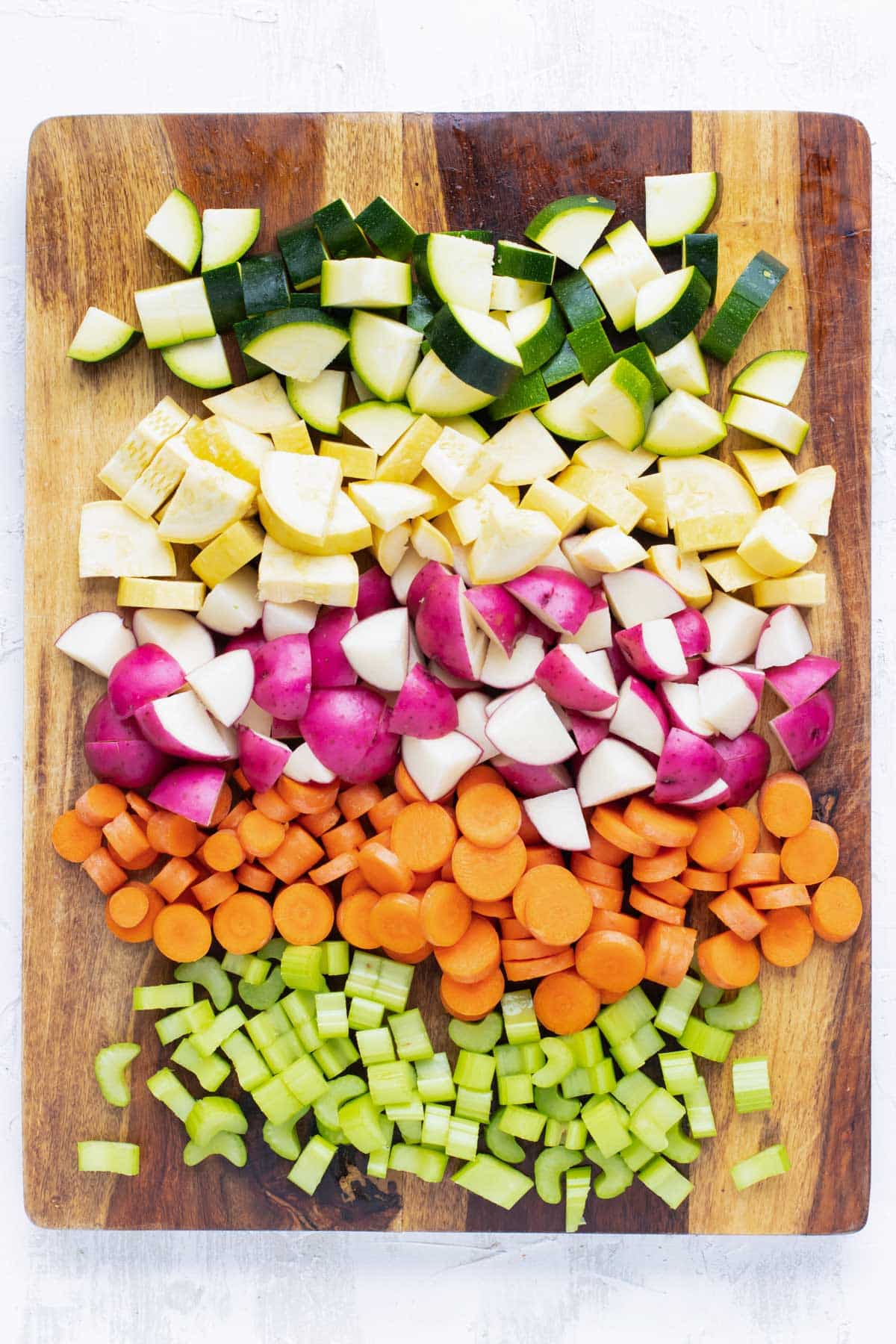 Chopped zucchini, yellow squash, red potatoes, carrots, and celery on a wooden cutting board.
