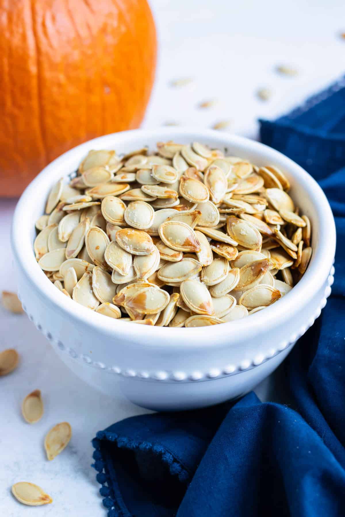Pumpkin seeds in a white bowl with a pumpkin behind it.