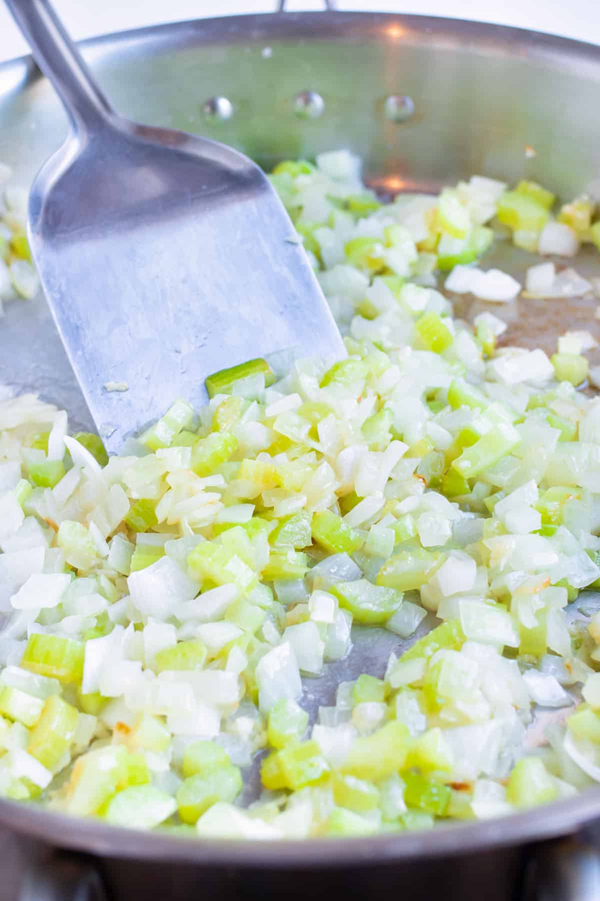 Diced onion and celery is sautéed in a skillet on the stove.
