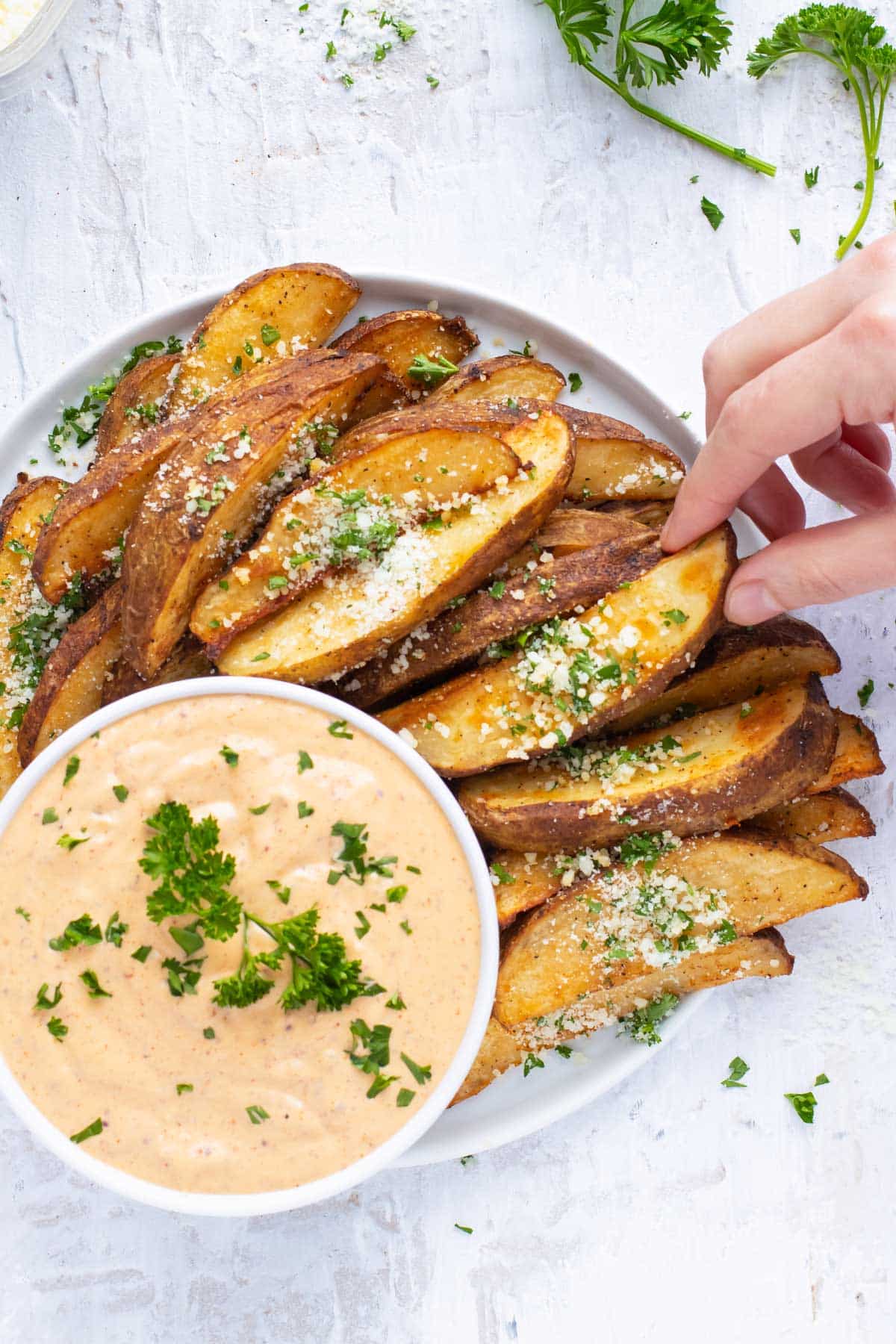 A hand grabbing a roasted potato wedge from a plate of them.