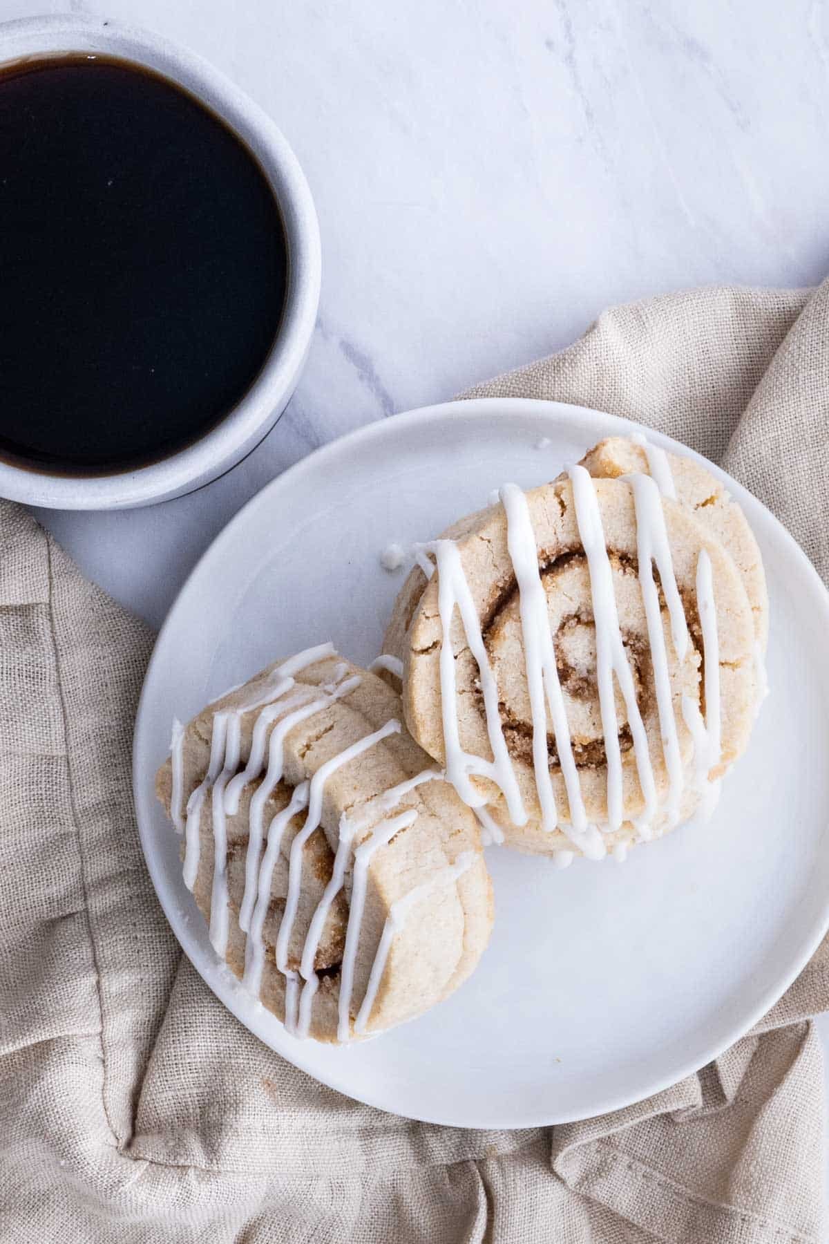 Two cinnamon Roll cookies are served on a white plate with coffee.