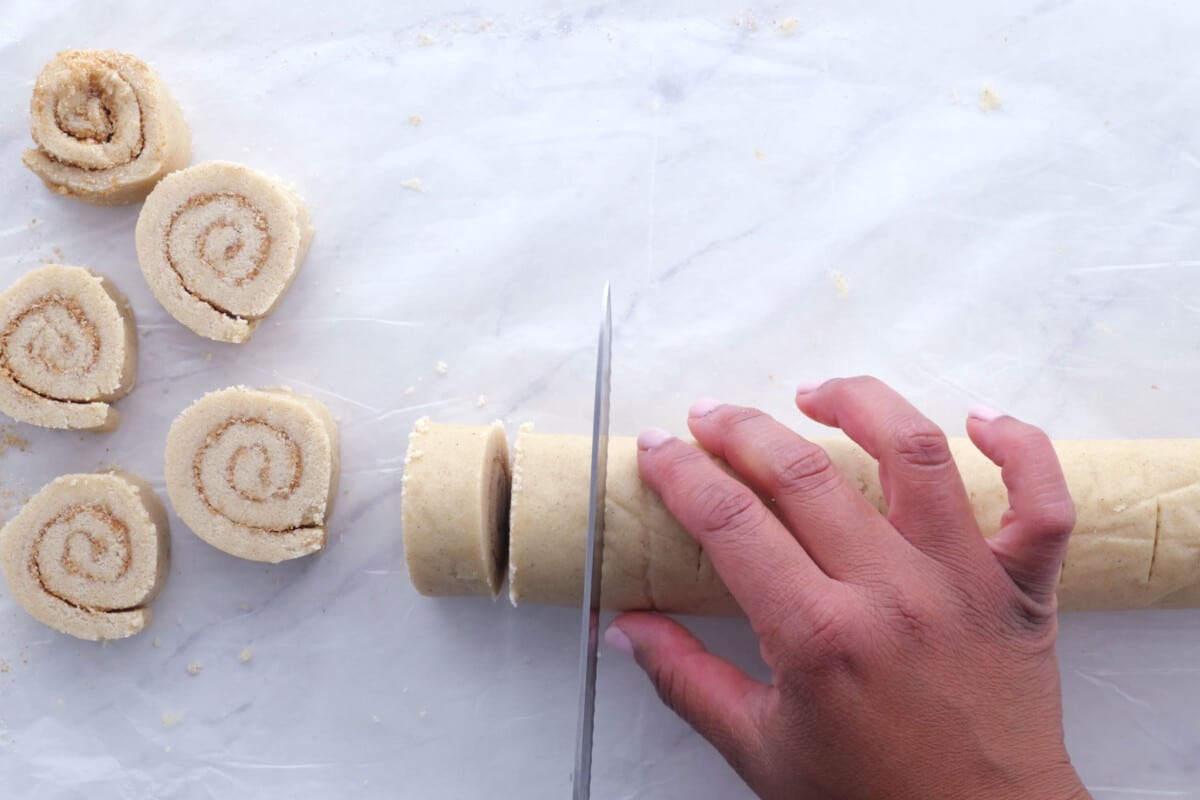 A knife slices the cinnamon roll cookie dough log.