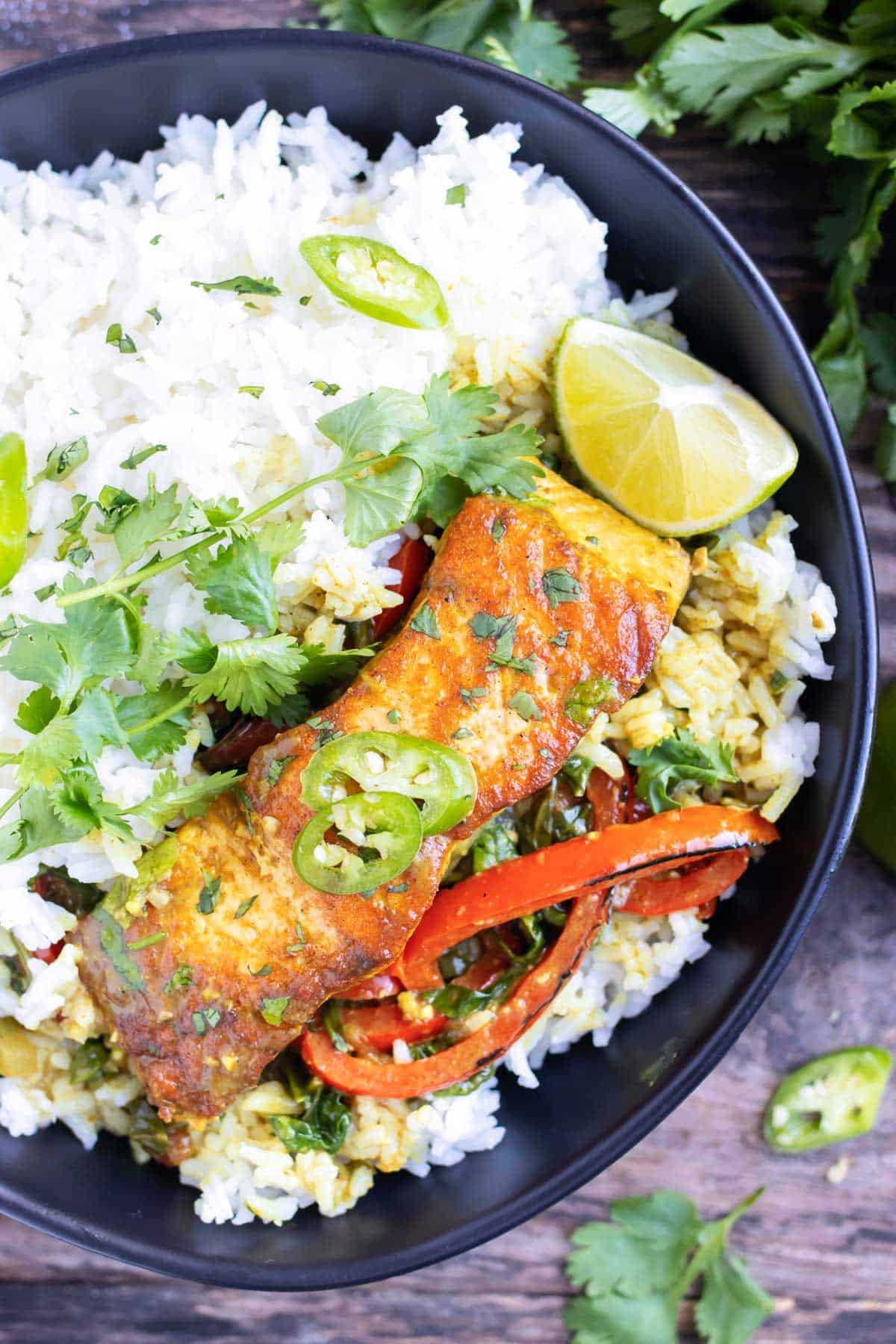 A bowl of salmon curry with coconut milk served over white rice with lime wedges and fresh cilantro.