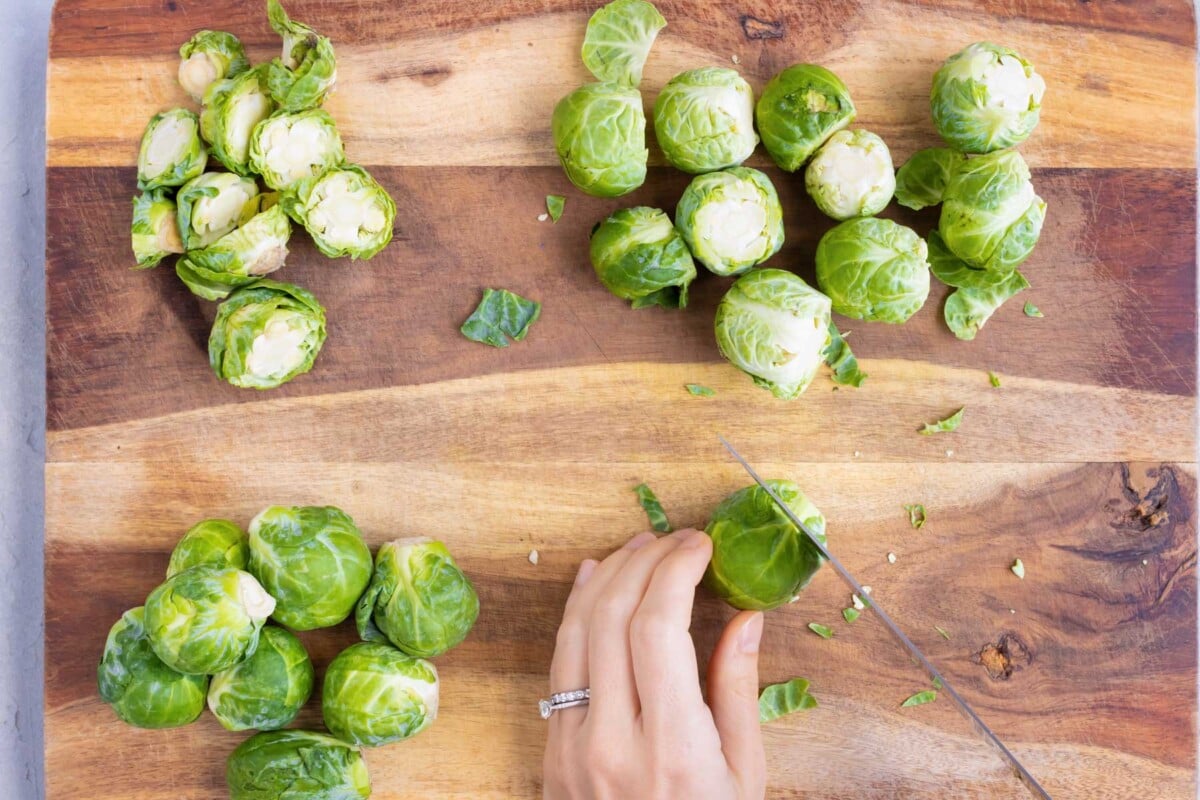 A wooden cutting board with Brussels sprouts that are getting the ends trimmed off with a knife.