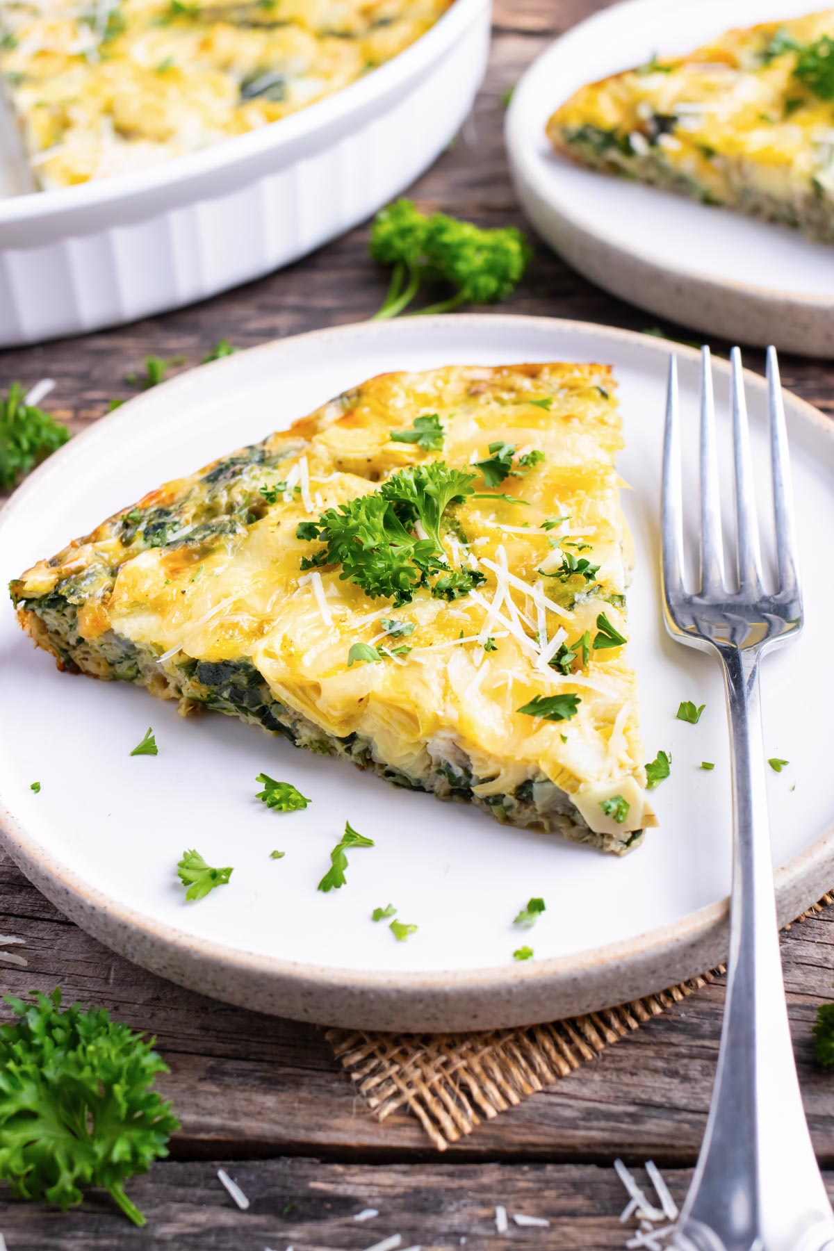 A white serving plate with a slice of spinach crustless quiche and a large baking dish full of it.