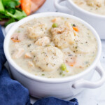 Chicken and dumpling soup in a white bowl with carrots and celery in the background.