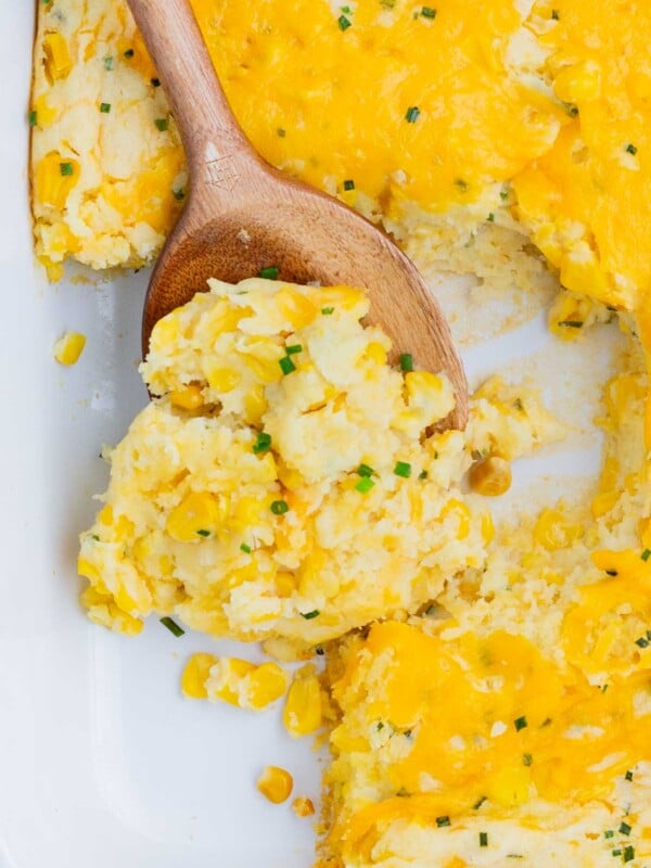 A wooden spoon scoops a serving of corn casserole from a pan.