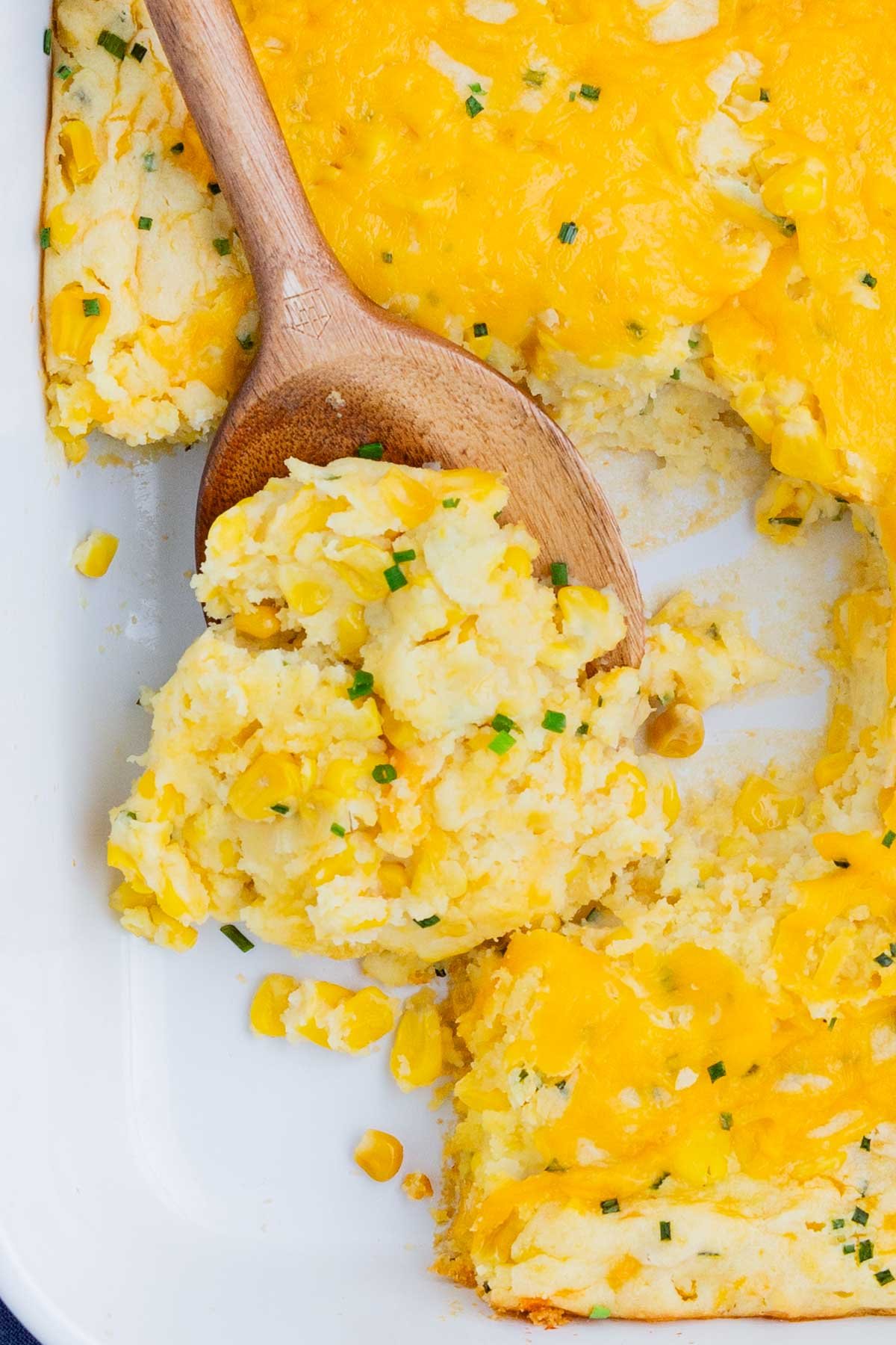 A wooden spoon scoops a serving of corn casserole from a pan.