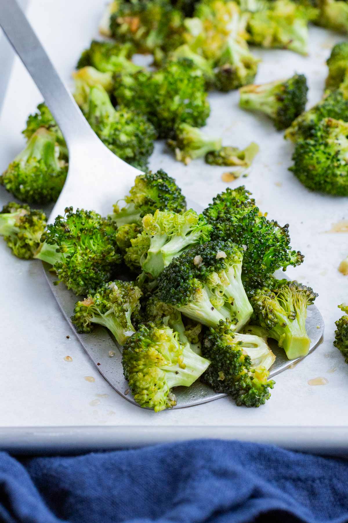 A metal spatula scoops roasted broccoli off of a tray.