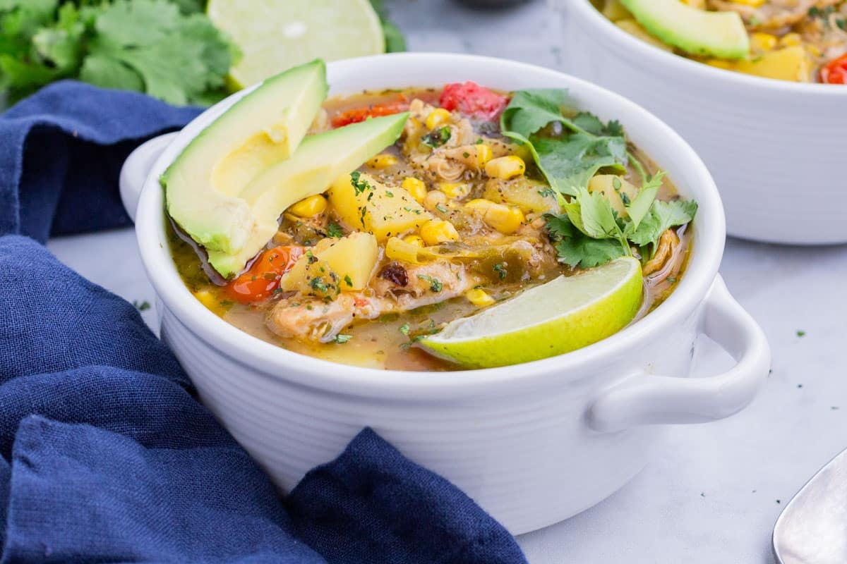 Two bowls of green chile chicken soup topped with avocado and lime wedges.