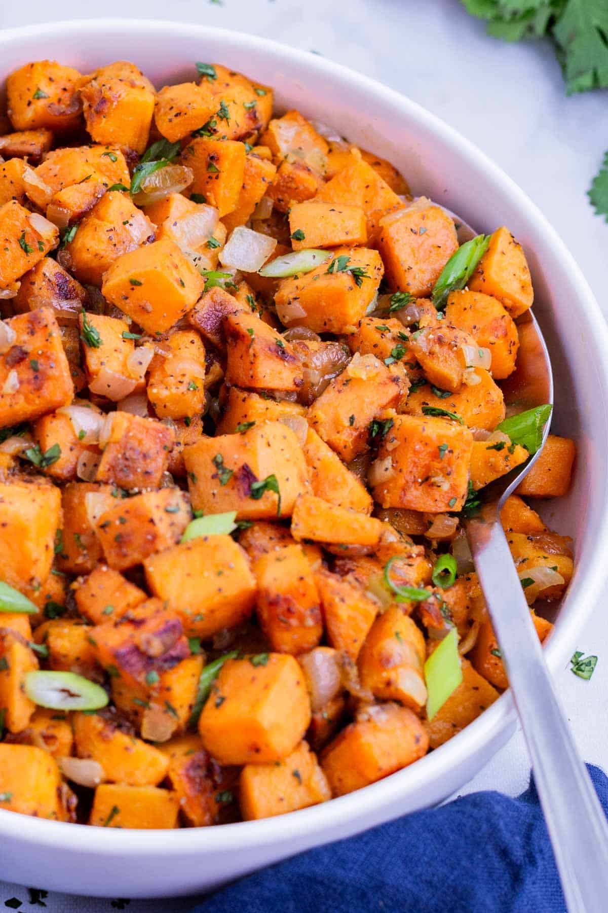 A metal spoons digs into a white bowl full of sauteed sweet potatoes.