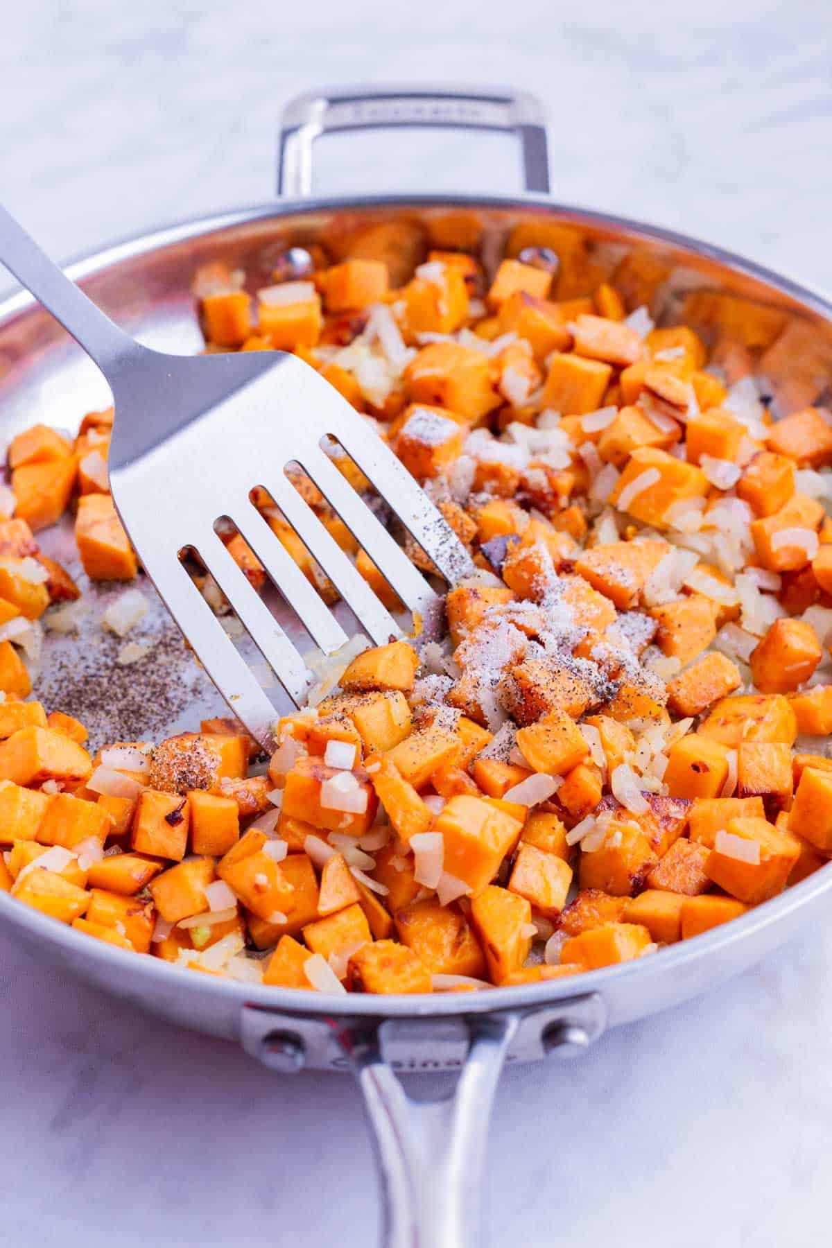 A spatula stirs sautéed sweet potatoes and onions together in a skillet.