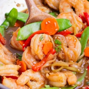 A wooden spoon stirs teriyaki shrimp stir fry in a wok.