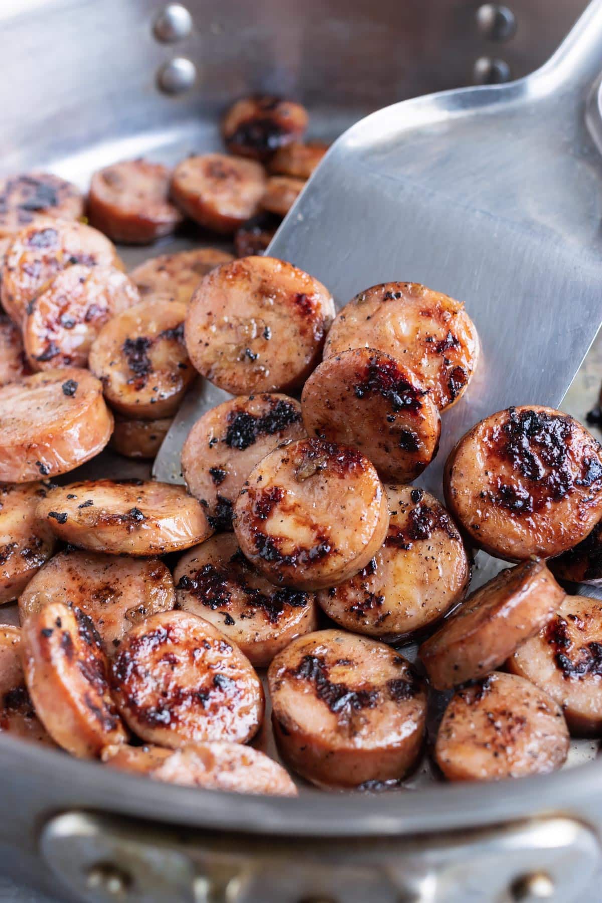 Chicken apple sausage that has been seared in a stainless steel skillet with a spatula.