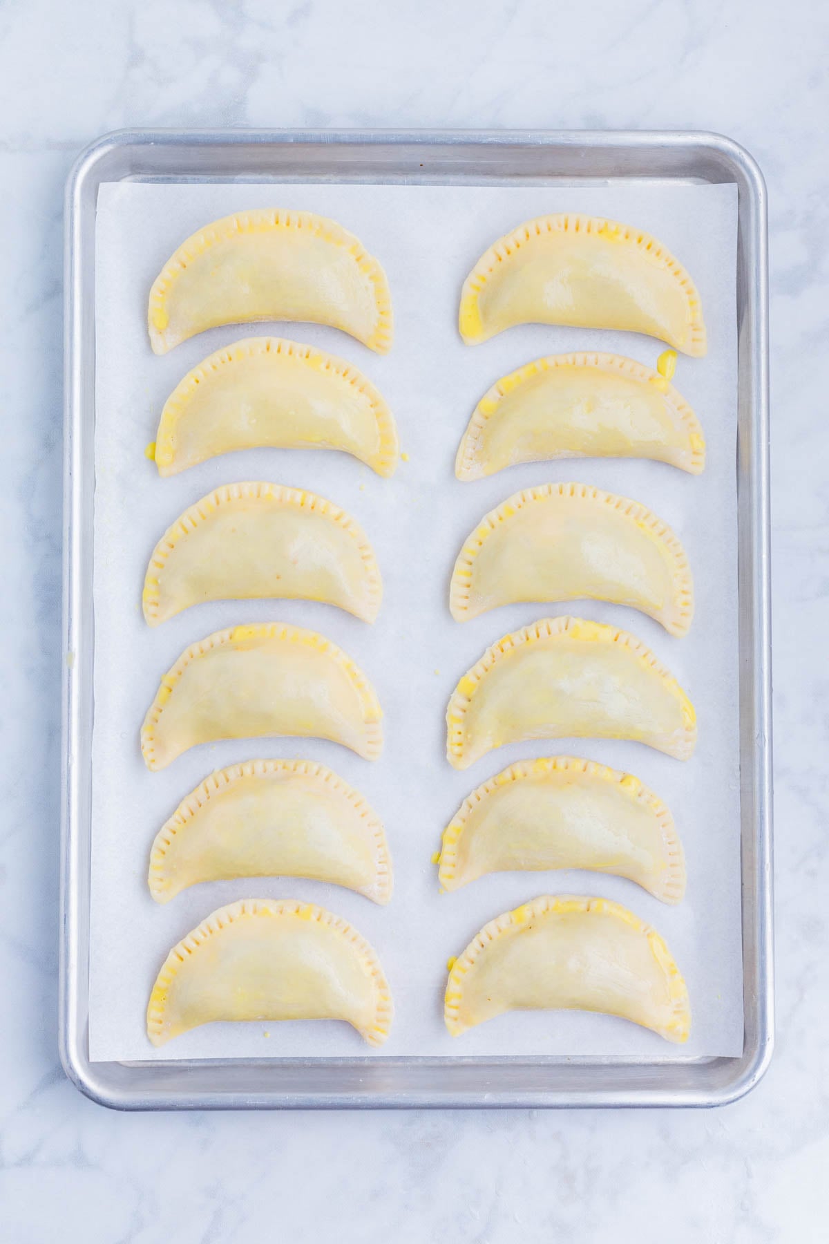 Twelve uncooked pie crust empanadas lined up on a parchment-lined sheet pan.