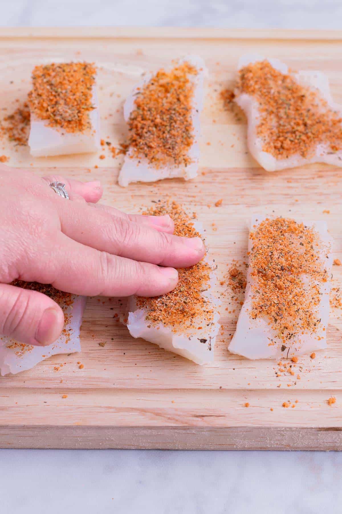 A hand presses blackened seasoning into fish filets.
