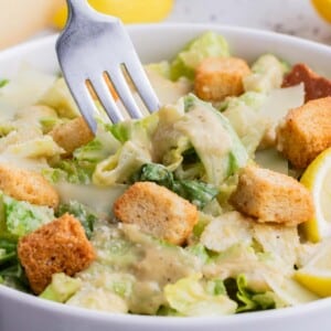 A fork digs into romaine lettuce with Caesar salad dressing in a white bowl.