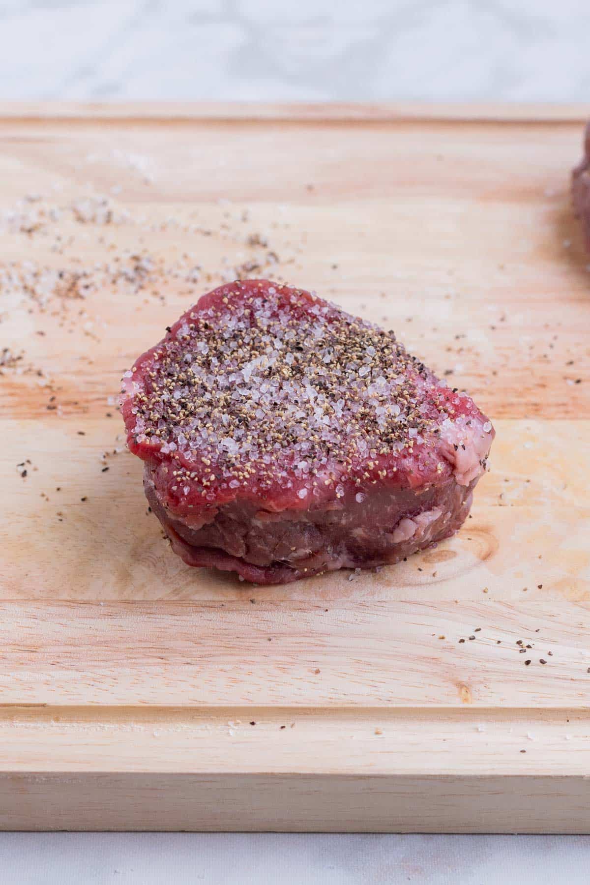 A filet mignon liberally seasoned with salt and pepper rests on a wooden cutting board.