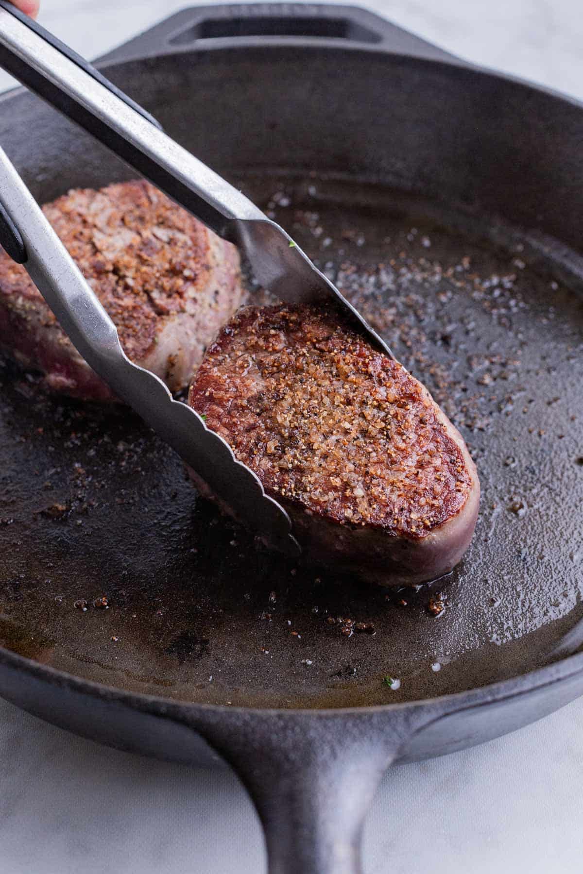 A pair of tongs flips a filet mignon in a cast-iron skillet.