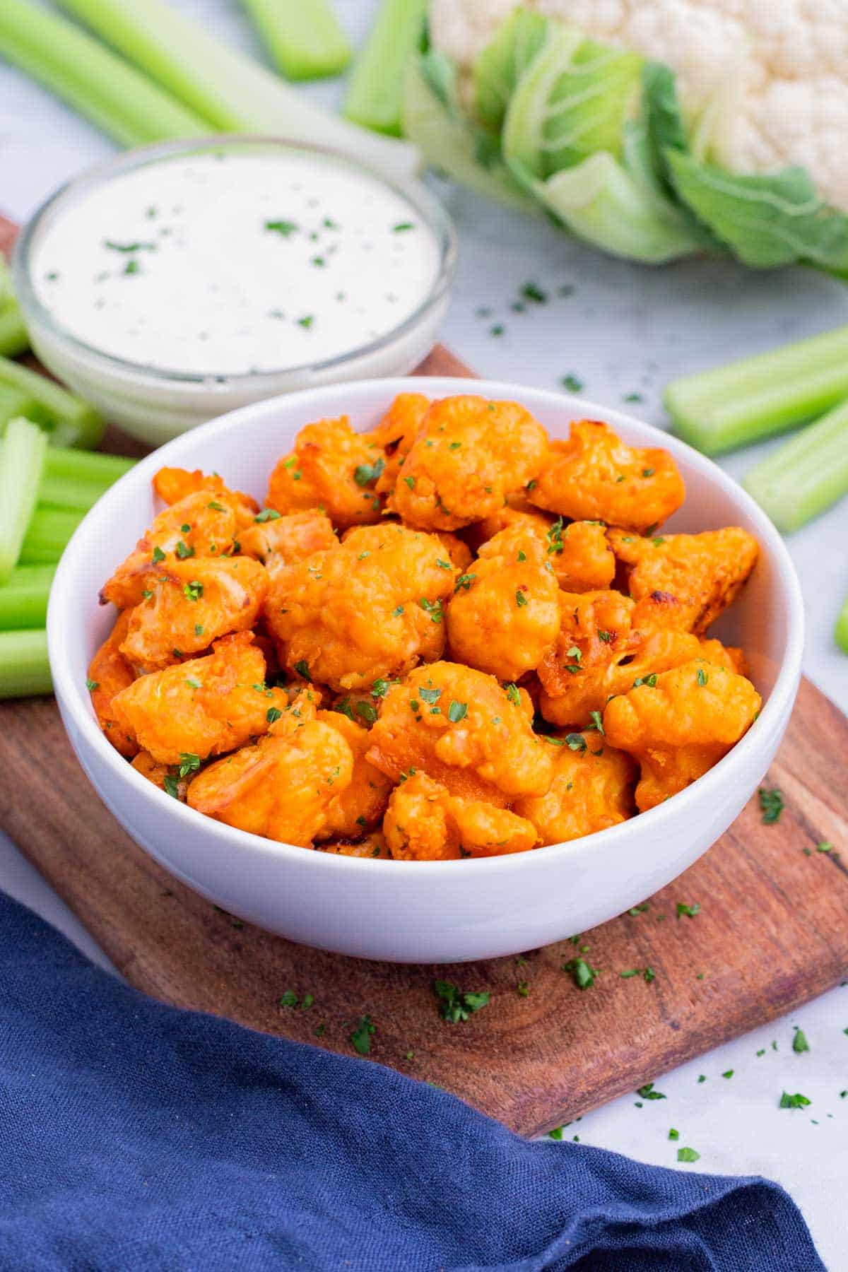 A head of cauliflower next to a serving platter of buffalo cauliflower bites, celery, and ranch dressing for a football party appetizer.