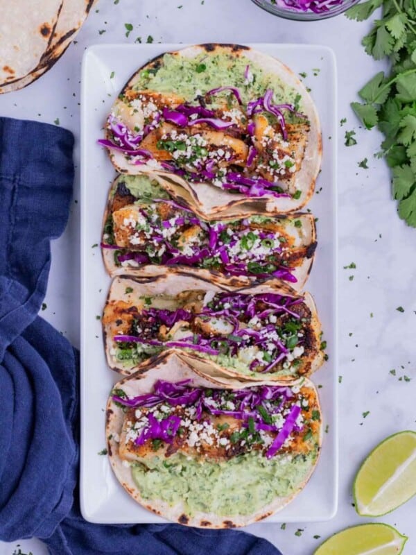 Four blackened fish tacos with avocado sauce and cabbage on a white plate.