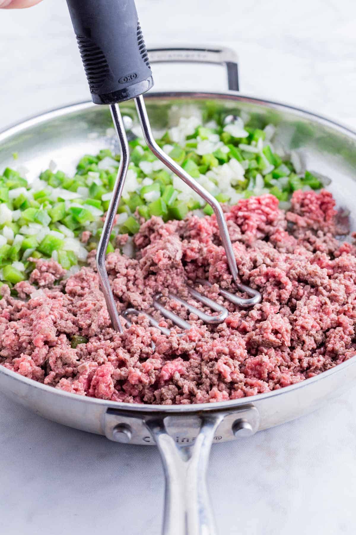 A potato masher breaks up ground beef in a skillet with veggies.