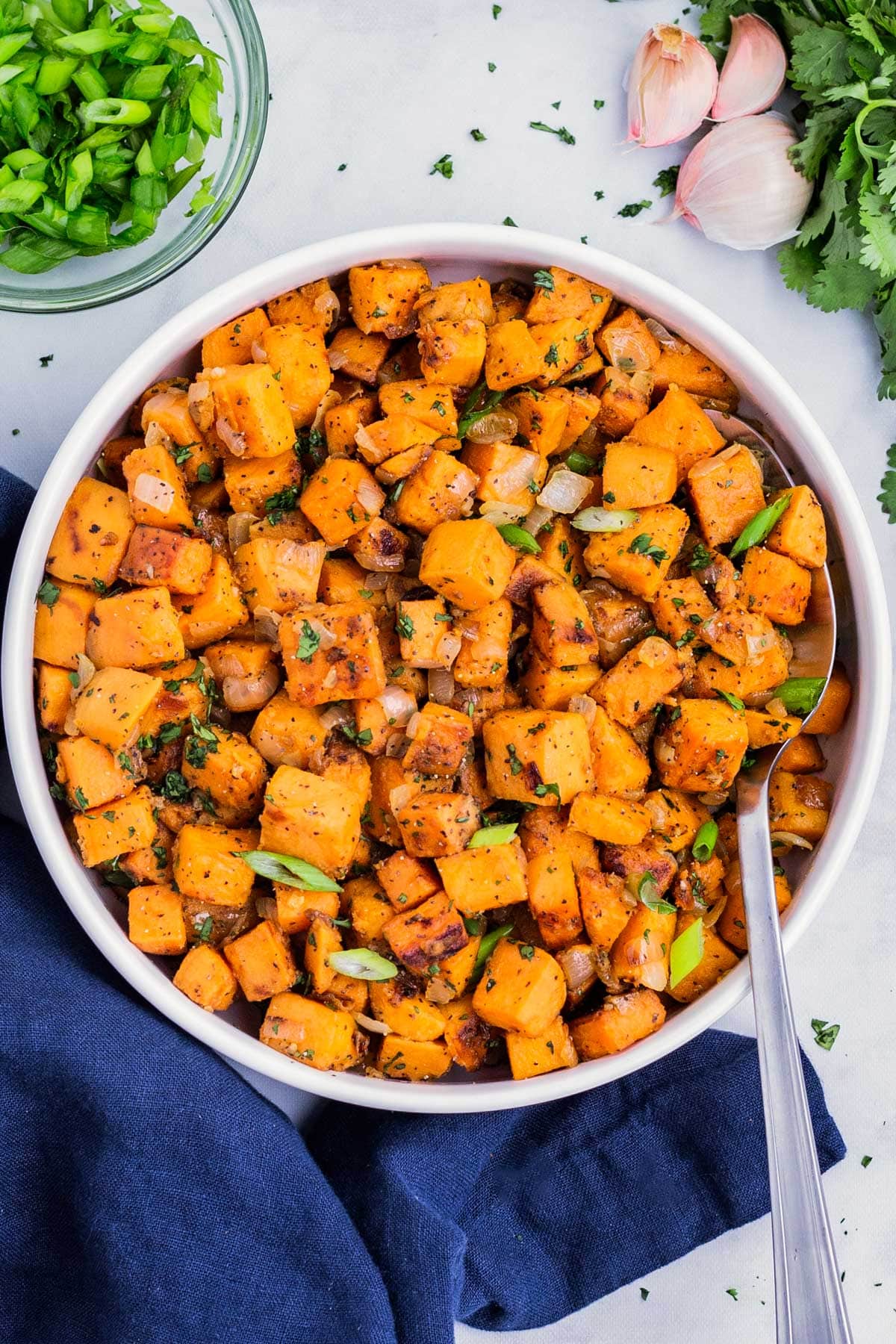 A white bowl is filled with diced sweet potatoes that have been cooked in a skillet and served with green onions.