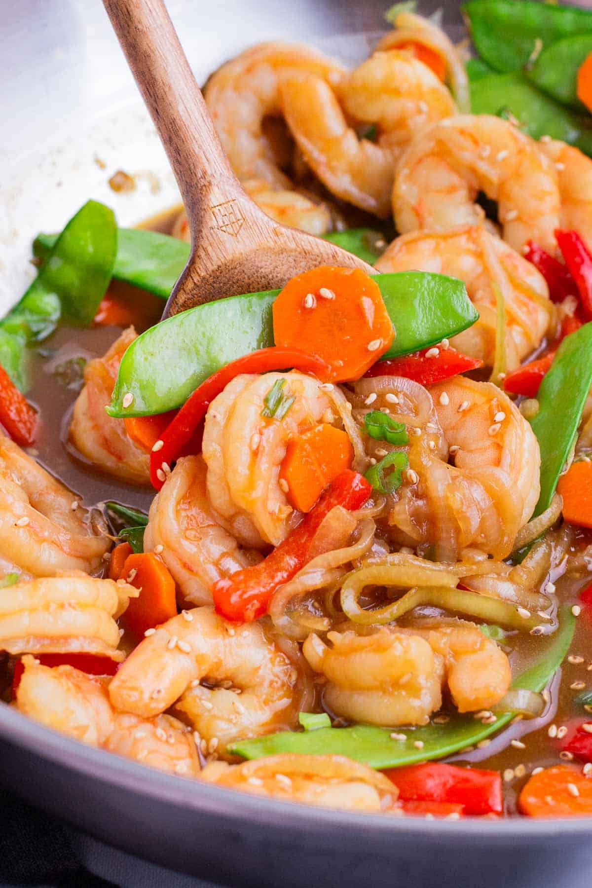 A wooden spoon stirs teriyaki shrimp stir fry in a wok.