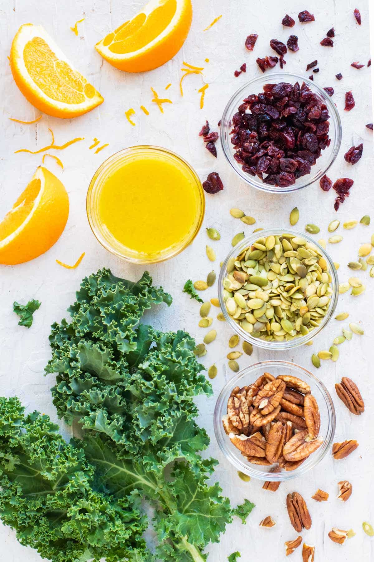 Oranges, cranberries, pumpkin seeds, and pecans in clear bowls with a bunch of kale.