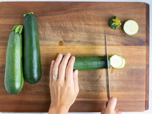 Three zucchinis are being sliced on a wooden cutting board.