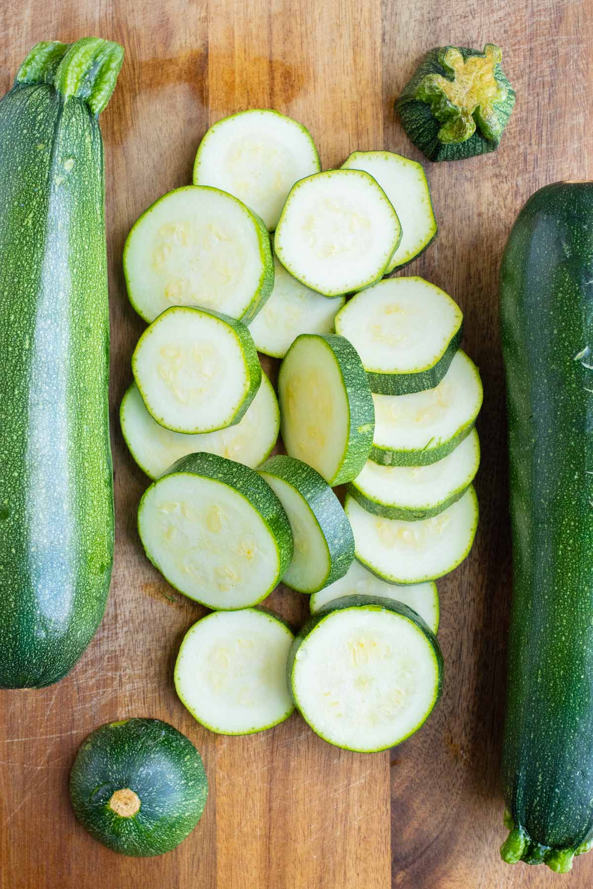 Zucchini is cut into thick slices on a cutting board with whole zucchinis next to them as preparation to freeze zucchini.