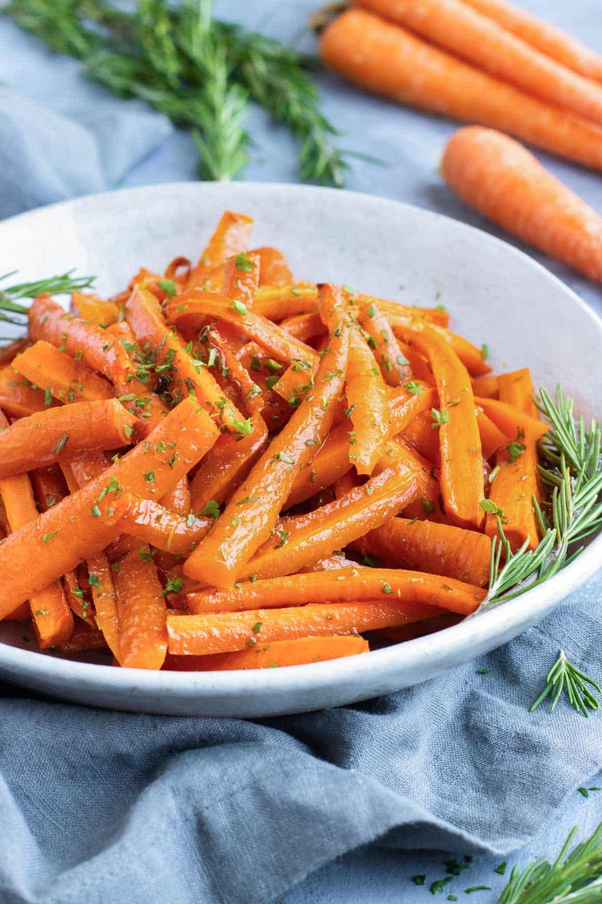 Herb roasted carrots in a white bowl with fresh rosemary and a blue linen napkin.