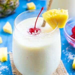 A frozen pina colada recipe in a clear glass with a pineapple wedge and a maraschino cherry.