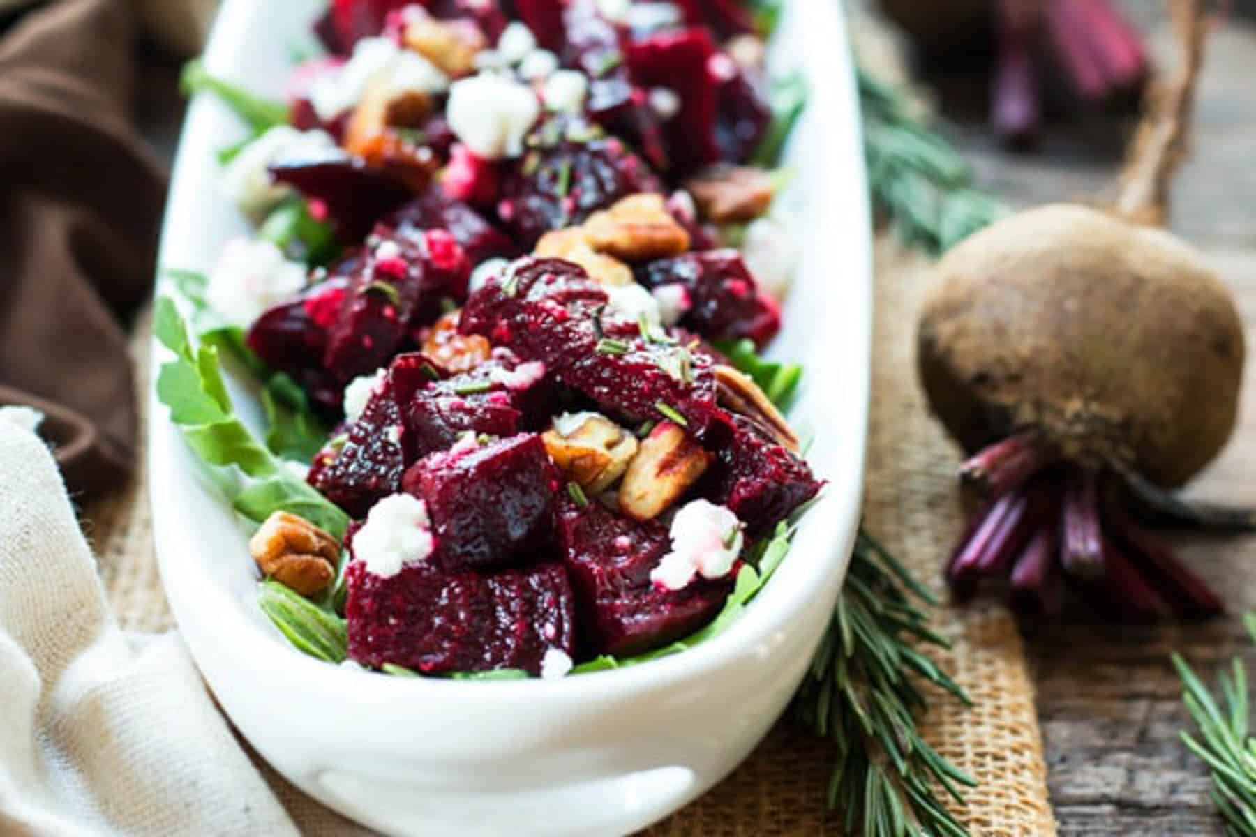 An easy beet and goat cheese side dish on a long white platter next to a whole raw beet and fresh rosemary.