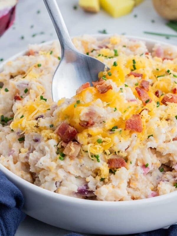 Mashed potatoes with bacon and cheese are served in a white bowl with a spoon scooping some up.