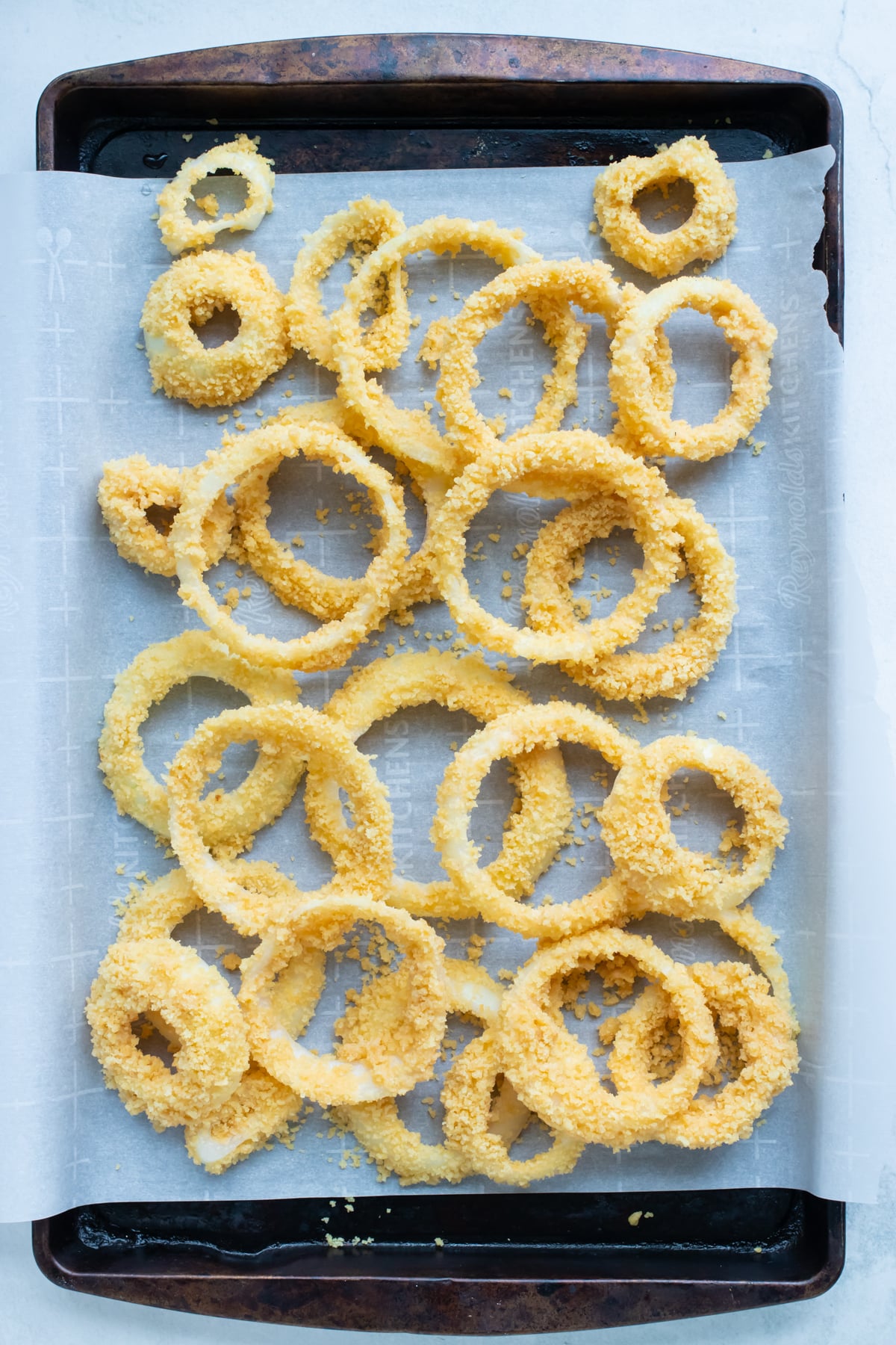 Breaded onion rings lined up on a parchment-lined sheet pan before going into the air fryer.