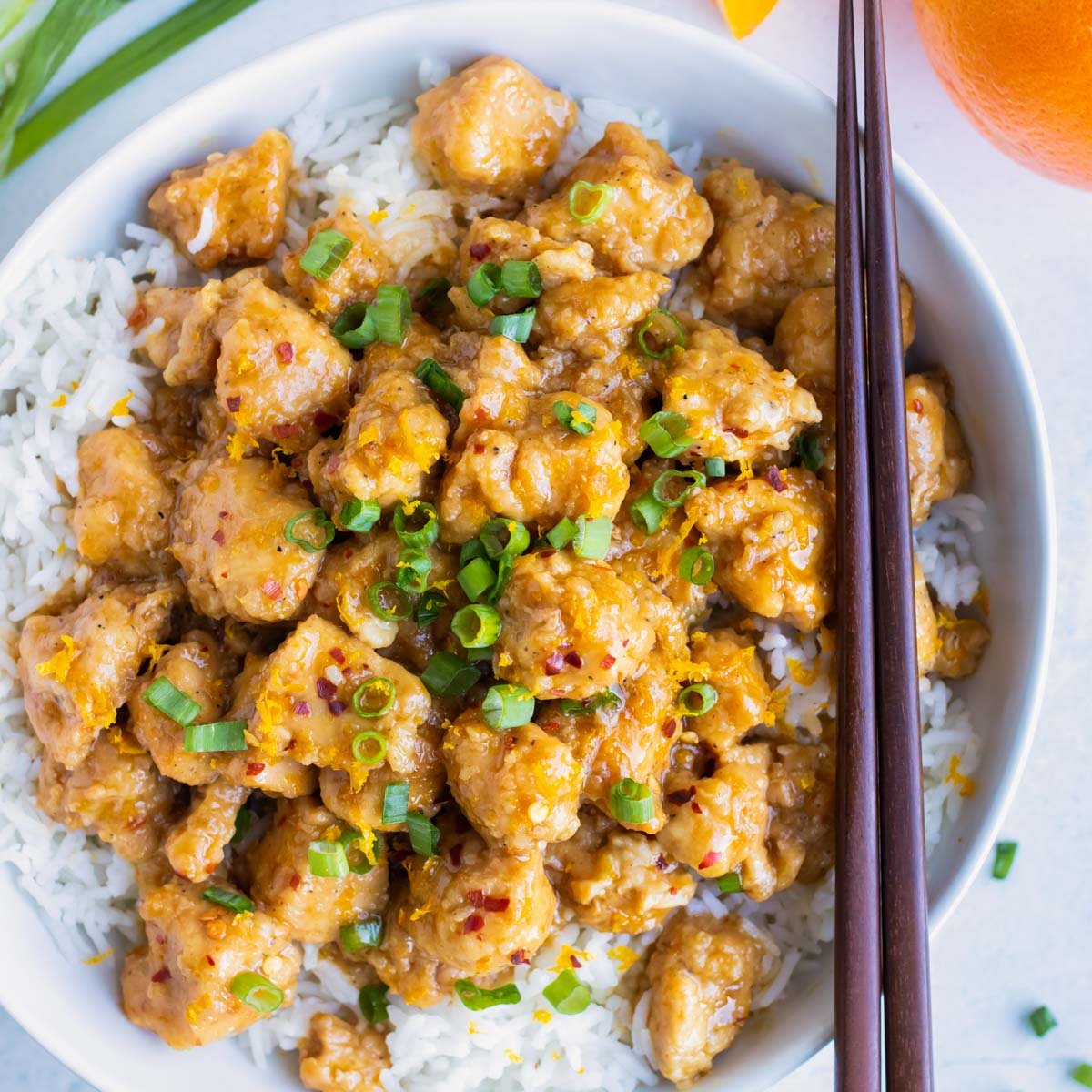 Healthy orange chicken with green onions and chopsticks in a white dinner bowl.