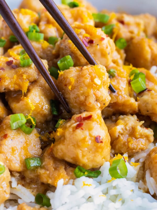 Chopsticks picking up a cube of orange chicken from a plate.