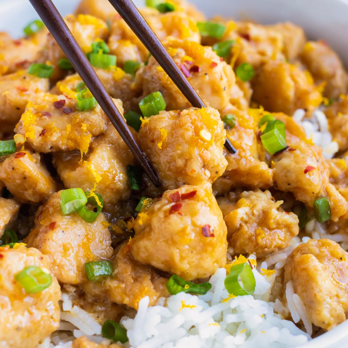 Chopsticks picking up a cube of orange chicken from a plate.