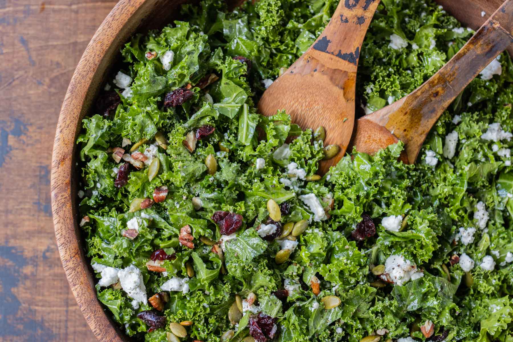 An overhead view of a chopped kale salad with pepitas and dried cranberries in a wooden bowl with two serving spoons.