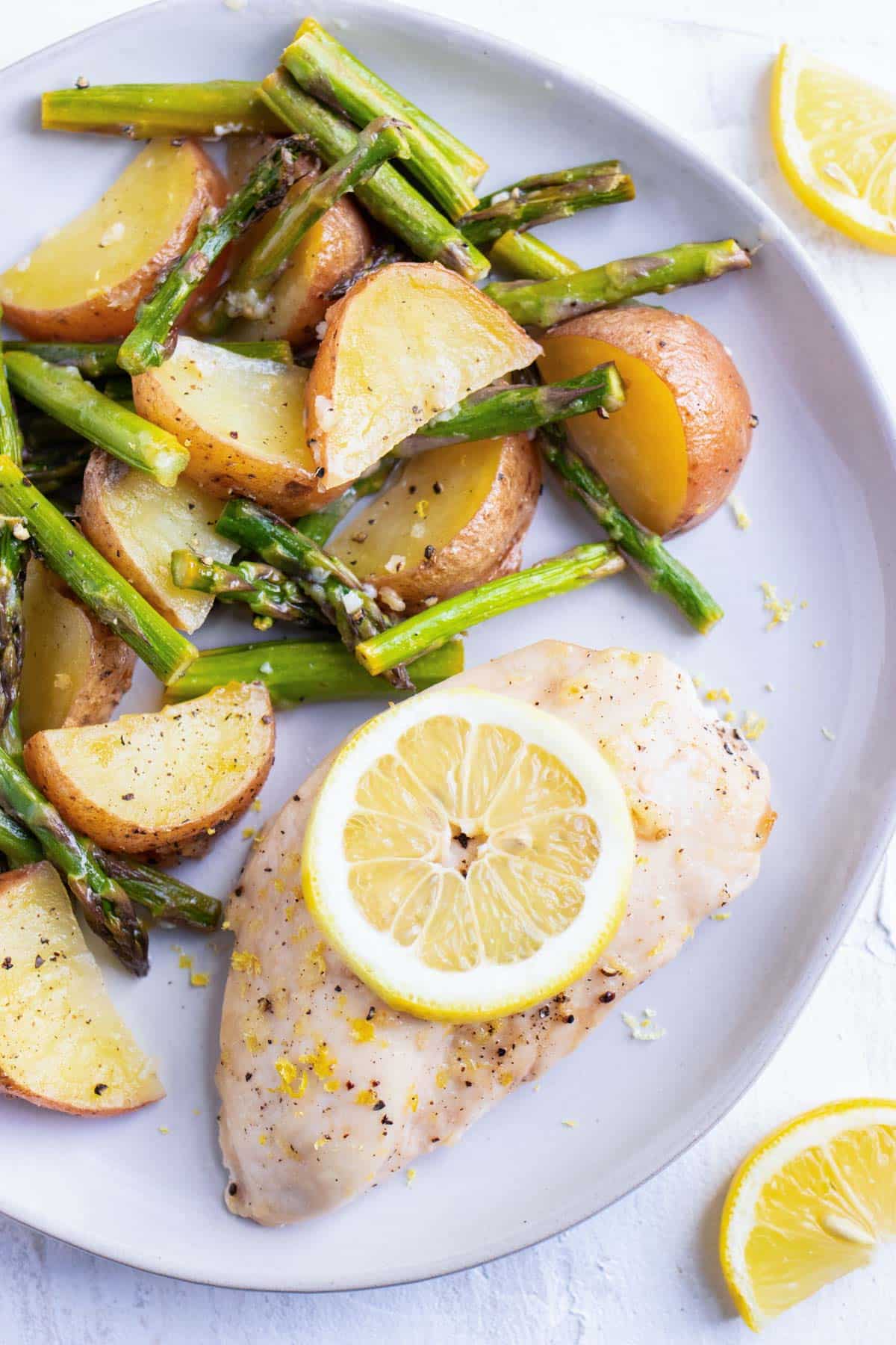 A single chicken breast with a lemon slice on top next to potato wedges and asparagus.