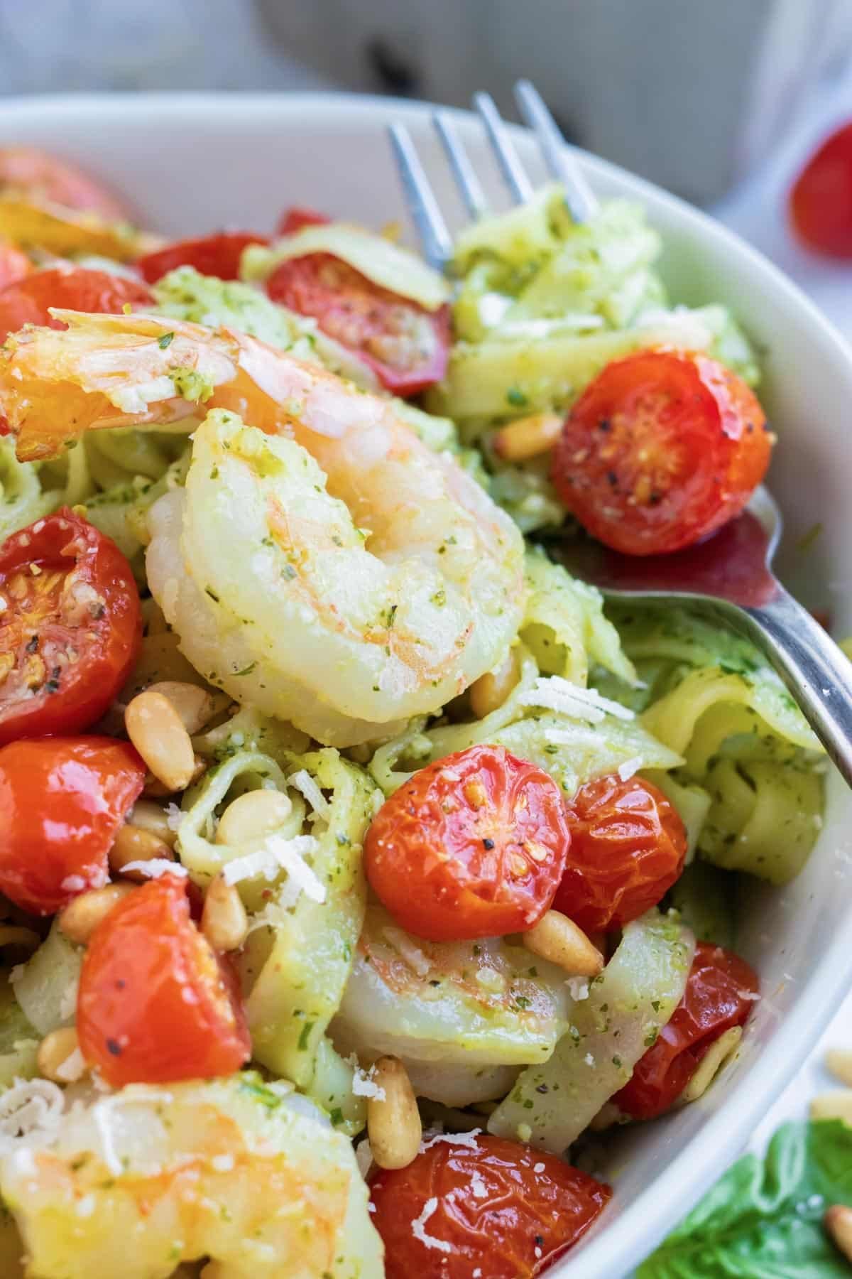 Shrimp pesto pasta in a white bowl with roasted cherry tomatoes and pine nuts.