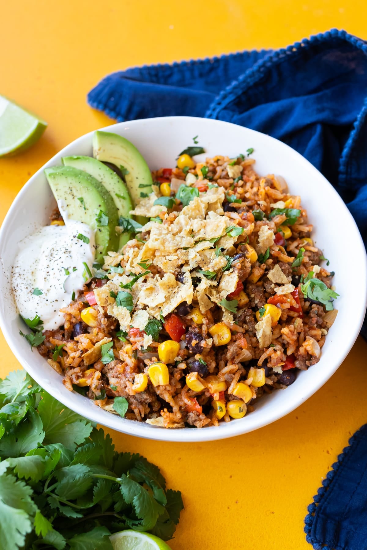 A Mexican beef and rice skillet recipe is served in a white bowl topped with avocado slices, sour cream, and crushed tortilla chips.