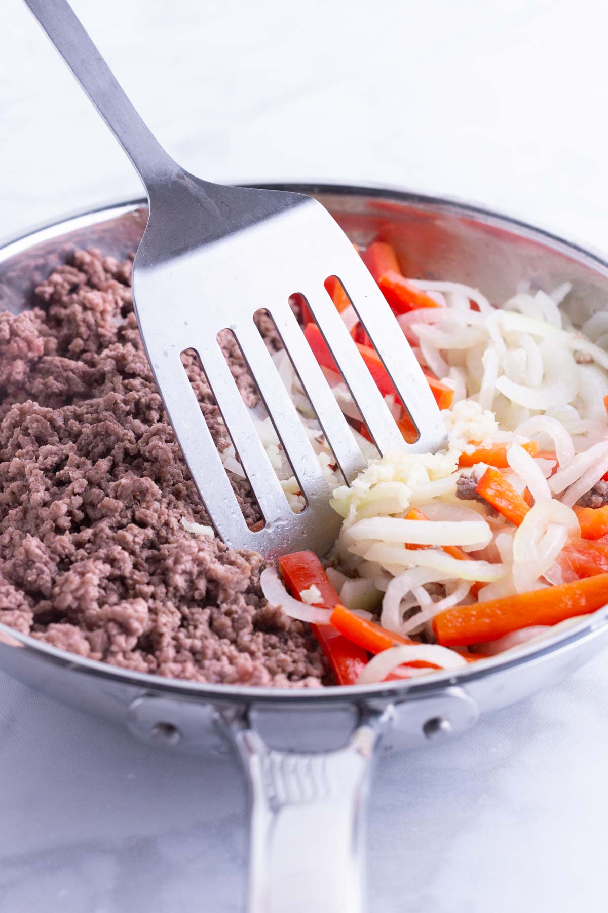 Onion and bell peppers are added to the beef in the skillet.