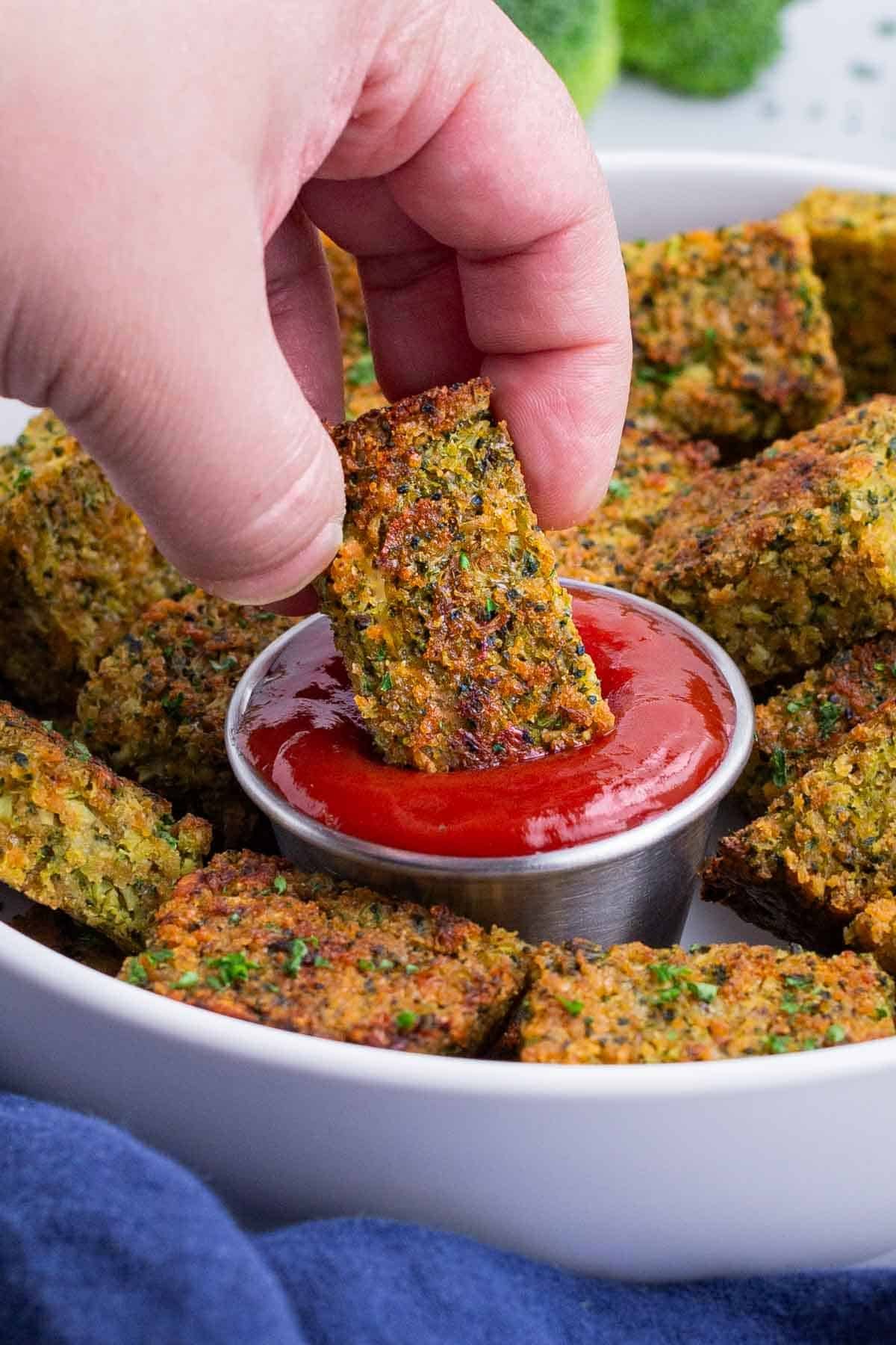 A hand dips a homemade broccoli tot into a bowl of ketchup.