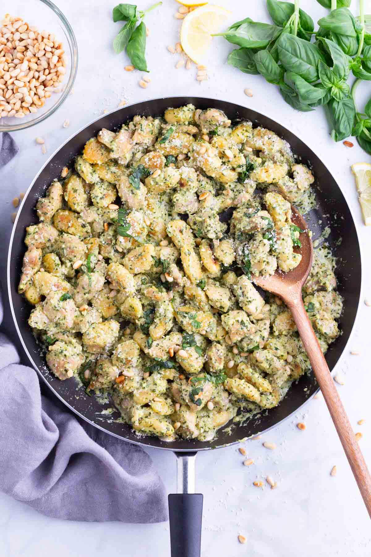 A wooden spoon is used to serve creamy pesto chicken gnocchi from a skillet.