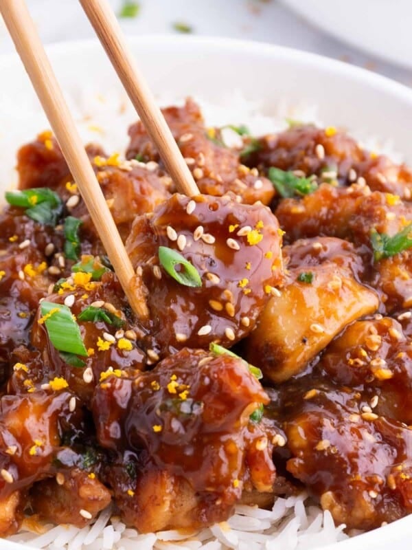 Chopsticks pick up a single piece of orange chicken from a bowl with rice.